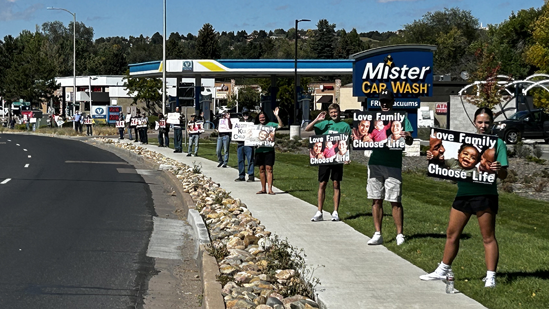 Group of protesters lined up on a sidewalk outside a Mister Car Wash, holding signs that say 'Love Family Choose Life'.